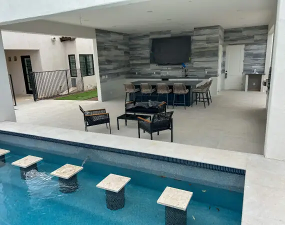 Outdoor kitchen and seating area with a large dining table, wicker chairs, and modern appliances, adjacent to a pool with in-water bar stools under a covered patio.