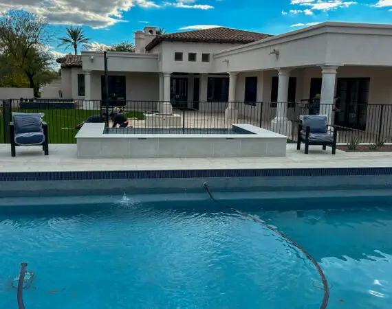 Beautiful backyard with a swimming pool and spa, featuring a fenced patio, cushioned chairs, green lawn, and a luxurious home with columns and tile roof.