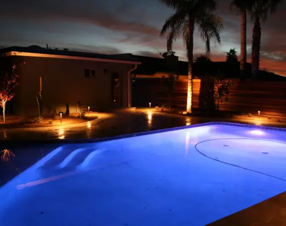 Evening view of a backyard with a glowing pool, surrounded by landscape lighting and palm trees, creating a serene and inviting atmosphere.