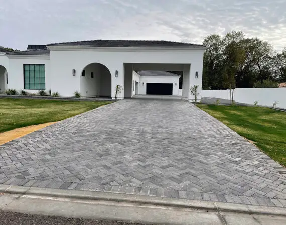 Elegant grey herringbone-patterned driveway leading to a modern white house with arches and a spacious front yard.